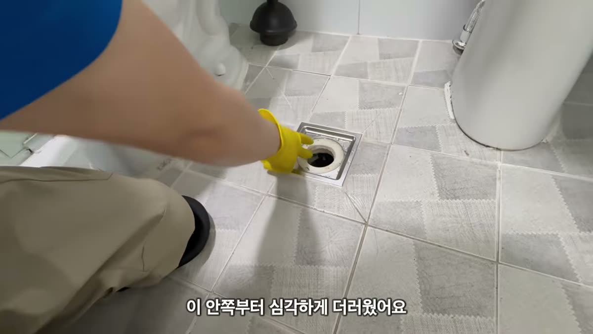 욕실 바닥 배수구 안쪽을 장갑 낀 손으로 닦는 장면